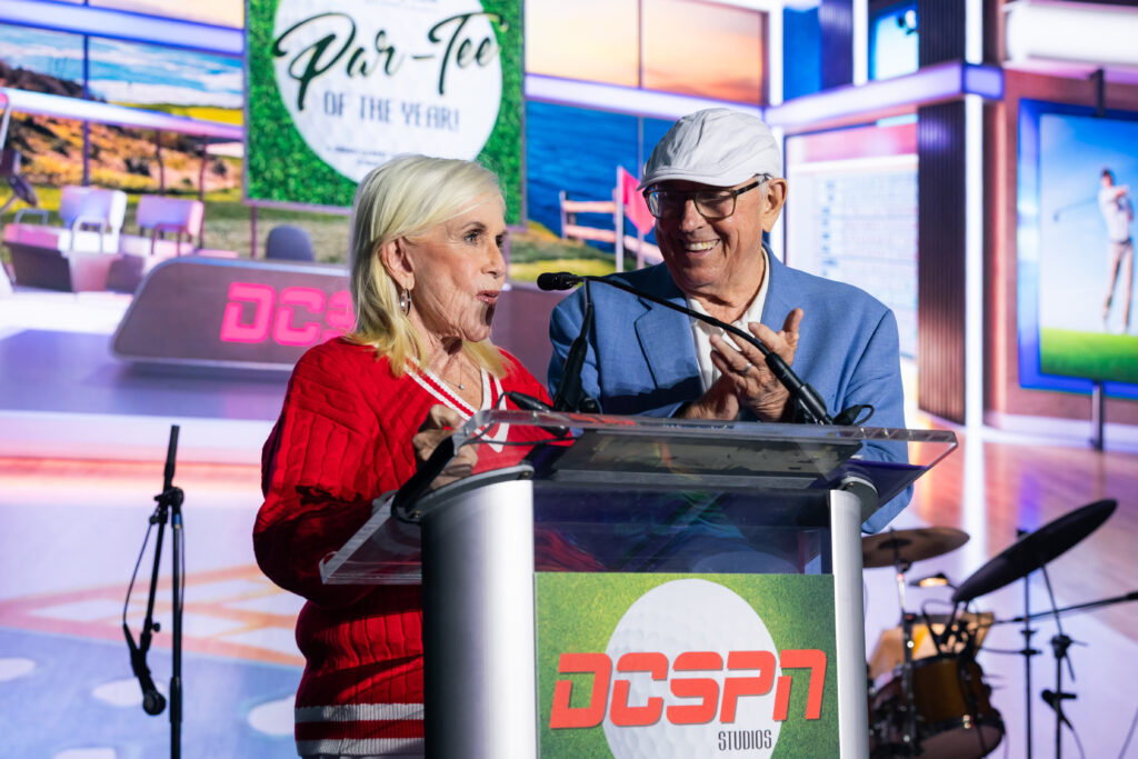 Two people standing on stage behind sports broadcaster table during event